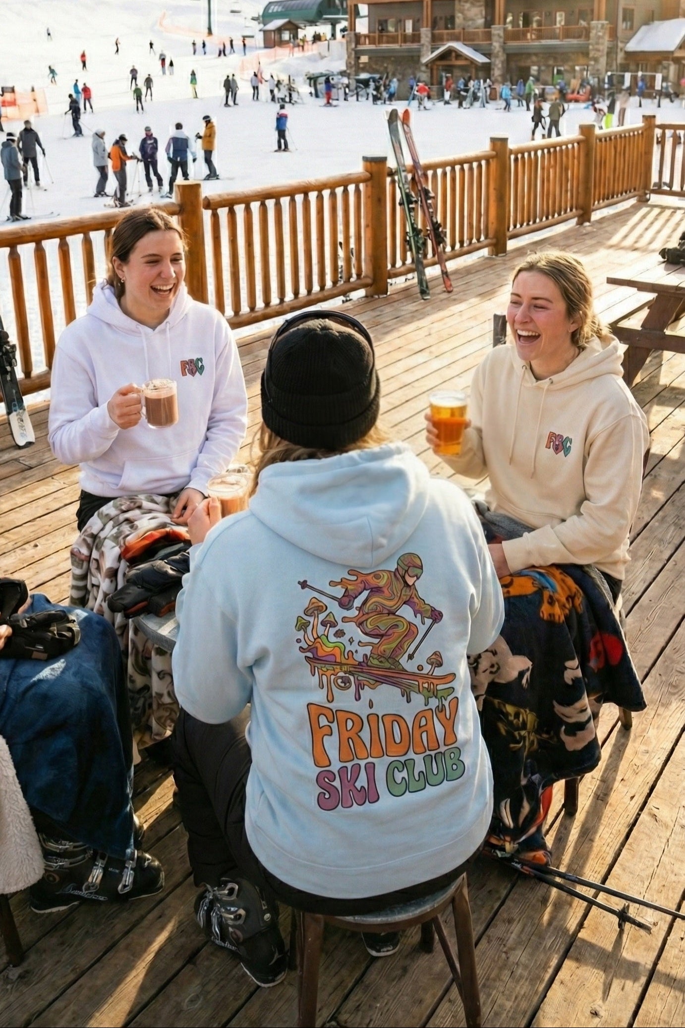 Three people sitting at a table with drinks on a wooden deck near a ski resort.